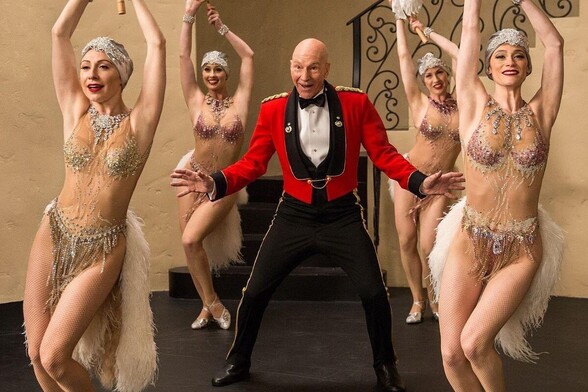 Sir Patrick Stewart wearing a fancy red jacket and black pants with gold piping, surrounded by dancing showgirls in slinky sequined costumes