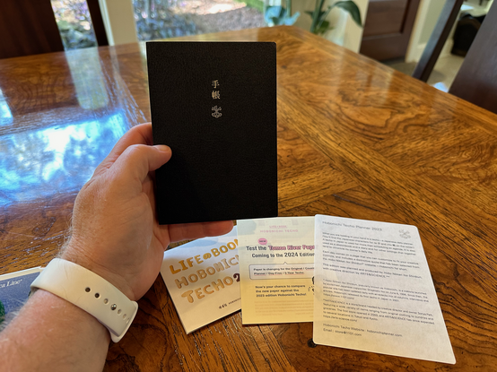 Mark’s hand, adorned with a white watch band and Apple Watch, holding a Hobonichi Techo — a life book. You can see the black textured cover stamped with the Japanese characters for Te and Cho in gold foil. It’s tiny and elegant, but still holds a whole year of pages.